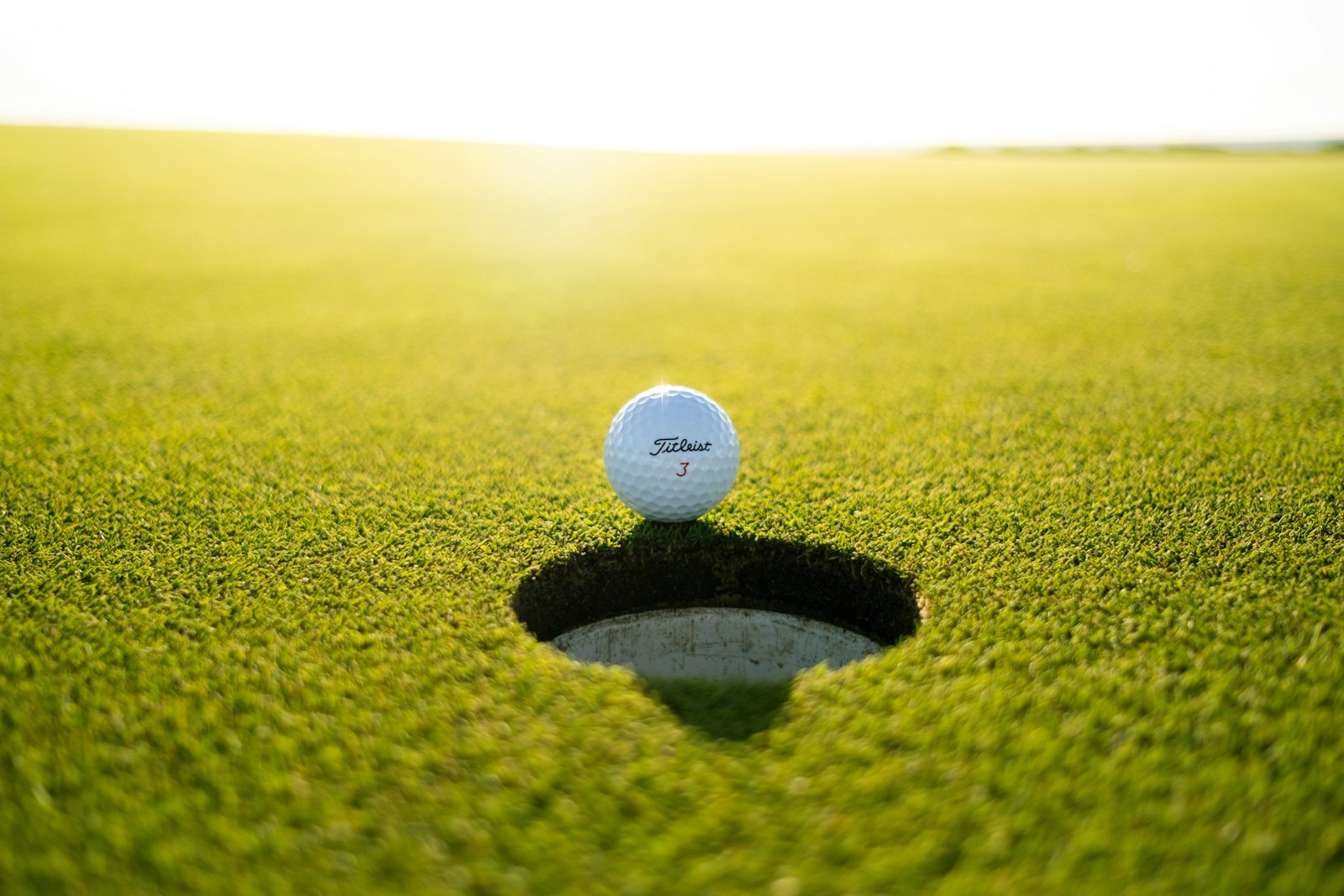 Golf ball resting at the edge of the hole on a green in low sun